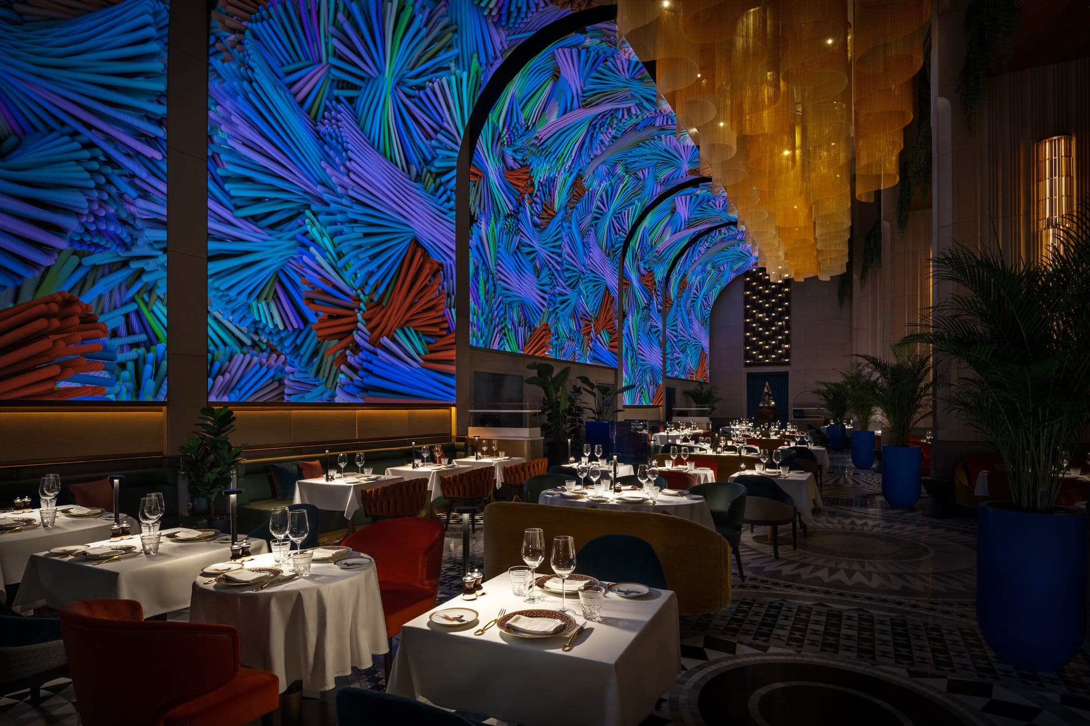 Tables laid with white linen at ZENON, under walls of immersive LED screens and a gold chain chandelier
