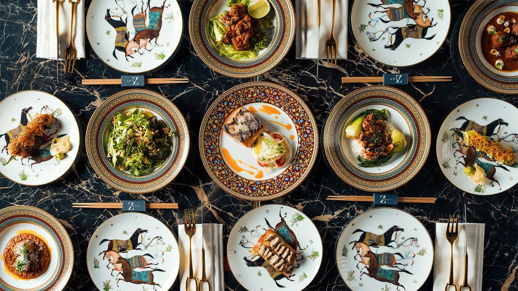 A table at ZENON seen from above, set with patterned porcelain, shared dishes and gold cutlery